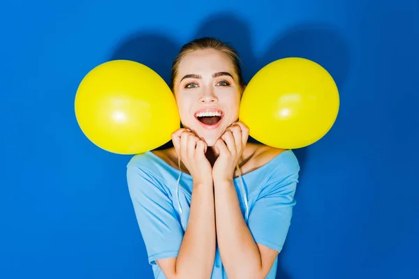 Muchacha joven atractiva sonriendo y sosteniendo globos por su cara sobre fondo azul - foto de stock