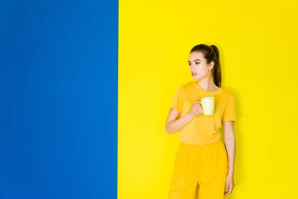 Weibliches Modell posiert mit Tasse in den Händen auf blauem und gelbem Hintergrund — Stockfoto