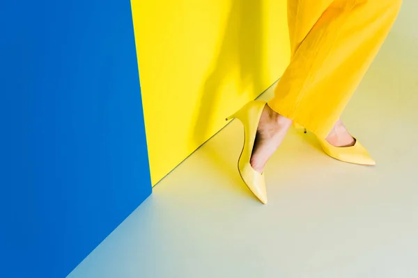 Vista recortada de pies femeninos en zapatos de tacón sobre fondo azul y amarillo - foto de stock