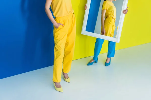 Vista recortada de mujeres probándose ropa frente al espejo sobre fondo azul y amarillo - foto de stock