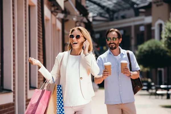 Mulher feliz falando por smartphone e segurando sacos de compras, homem carregando copos de café descartáveis na rua — Fotografia de Stock