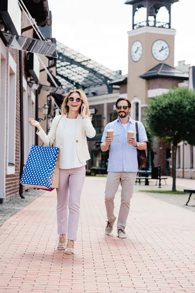 Esposa hablando por teléfono inteligente y sosteniendo bolsas de compras, marido llevando tazas de café desechables en la calle - foto de stock