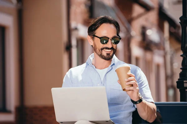 Bell'uomo sorridente seduto sulla panchina con computer portatile e caffè in tazza di carta — Foto stock