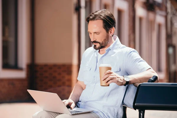 Homem bonito sentado no banco com laptop e copo de café descartável — Fotografia de Stock