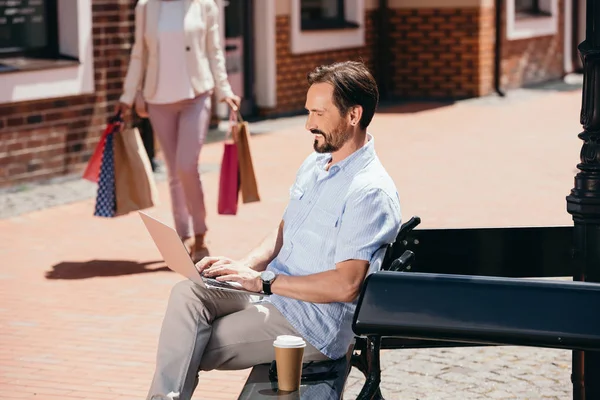 Hombre guapo sentado en el banco con el ordenador portátil, mujer caminando con bolsas de compras en la calle - foto de stock