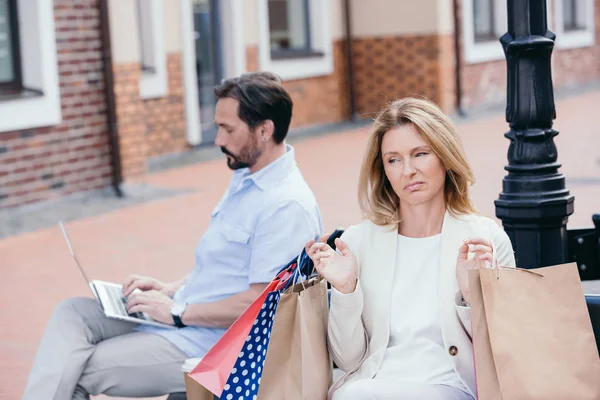 Man using laptop on bench, sad woman sitting with shopping bags — Stock Photo