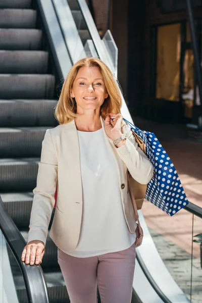 Mujer atractiva sonriente de pie en escaleras mecánicas con bolsas de compras en el centro comercial - foto de stock