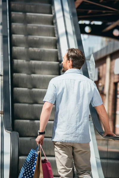 Back view of man standing on escalator with shopping bags in mall — Stock Photo