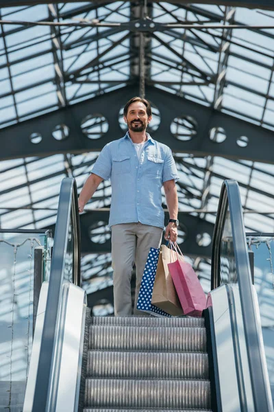 Vista de ángulo bajo del hombre guapo de pie en la escalera mecánica con bolsas de compras - foto de stock