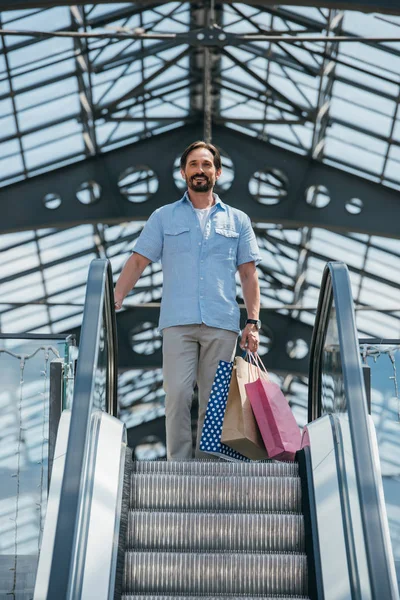 Vue à angle bas du bel homme debout sur l'escalator avec des sacs à provisions — Photo de stock