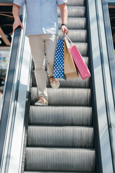 Imagen recortada de hombre caminando en escaleras mecánicas con nags de compras - foto de stock