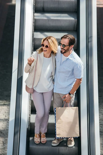 Vue grand angle du couple heureux dans les lunettes de soleil regardant loin sur l'escalator dans le centre commercial — Photo de stock