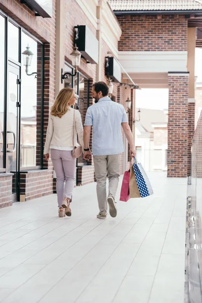 Vista trasera de pareja cogida de la mano y caminando con bolsas de compras - foto de stock