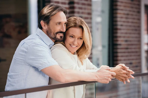 Feliz pareja abrazándose en el balcón y mirando hacia otro lado - foto de stock