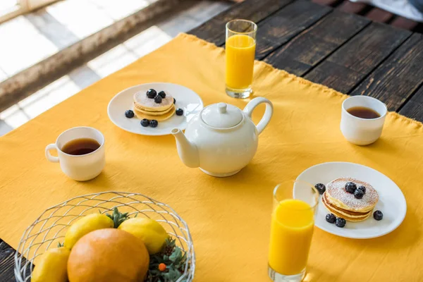 Vista da vicino di frittelle, tazze di tè e bicchieri di succo di frutta per la colazione sul tavolo con tovaglia gialla — Foto stock