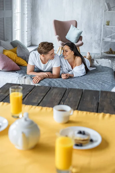 Foco seletivo do café da manhã na mesa e casal multirracial no amor deitado na cama no quarto — Fotografia de Stock