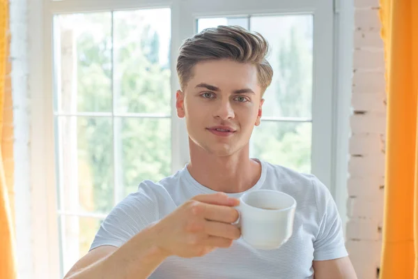 Retrato de hombre joven con taza de té mirando a la cámara en casa - foto de stock
