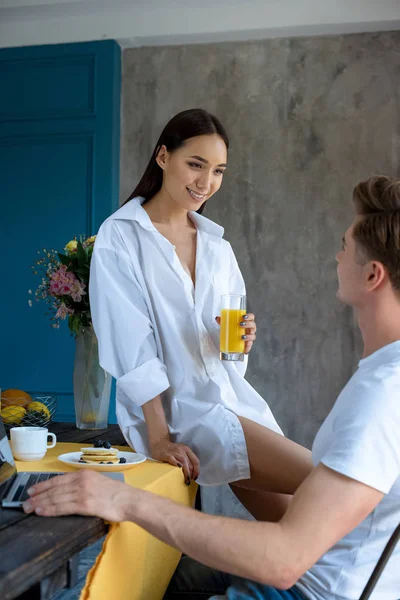 Smiling asian woman with glass of juice looking at boyfriend at table with laptop and breakfast at home — Stock Photo