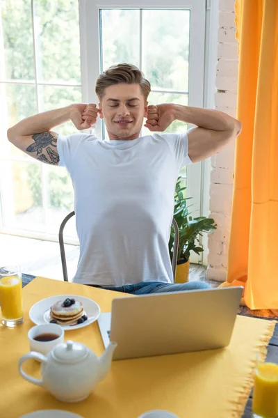 Portrait of young man stretching while sitting at table with breakfast and laptop at home — Stock Photo