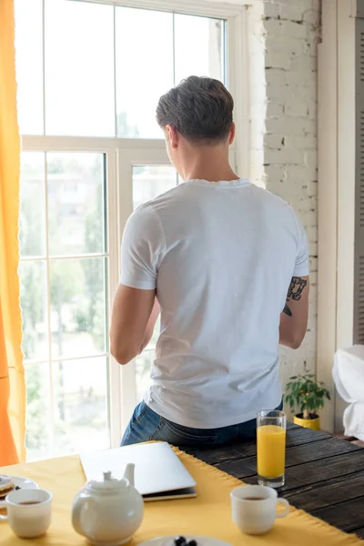 Visão traseira do homem de pé na janela com café da manhã na mesa em casa — Fotografia de Stock