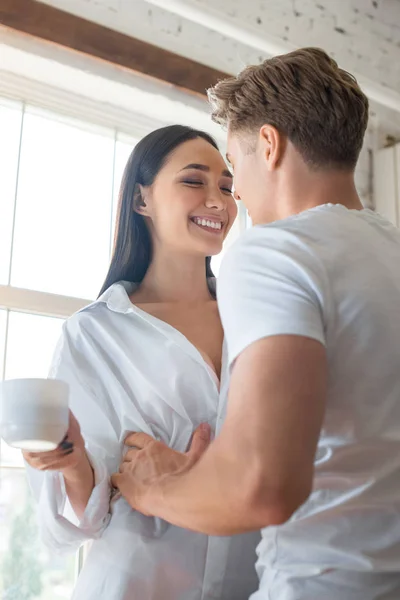 Caucasan boyfriend hugging smiling asian woman with cup of coffee at home — Stock Photo