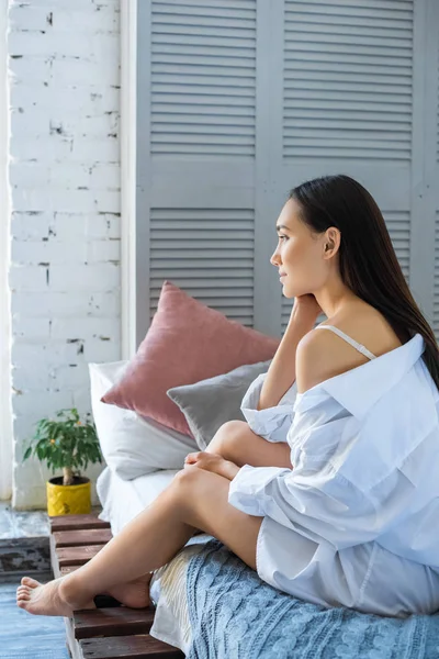 Side view of pensive asian woman in white shirt resting on bed at home — Stock Photo