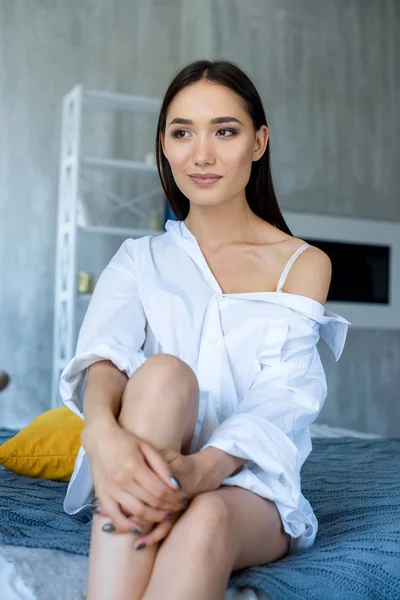 Retrato de mujer asiática pensativa en camisa blanca descansando en la cama en casa - foto de stock