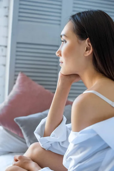 Side view of pensive asian woman in white shirt resting on bed at home — Stock Photo