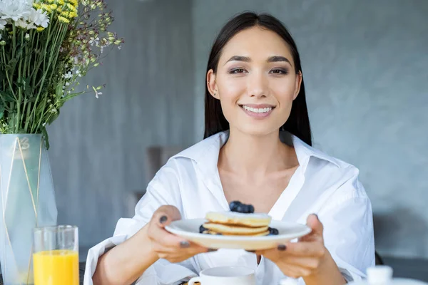 Retrato de mujer asiática sonriente con panqueques servidos con arándanos en el plato en las manos en casa - foto de stock