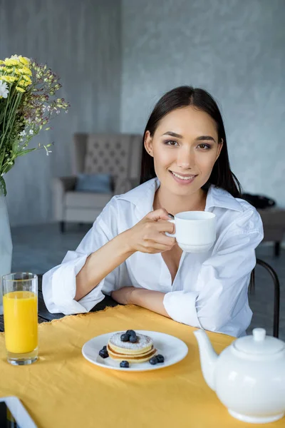 Portrait of smiling asian woman with cup of tea at table with breakfast at home — Stock Photo