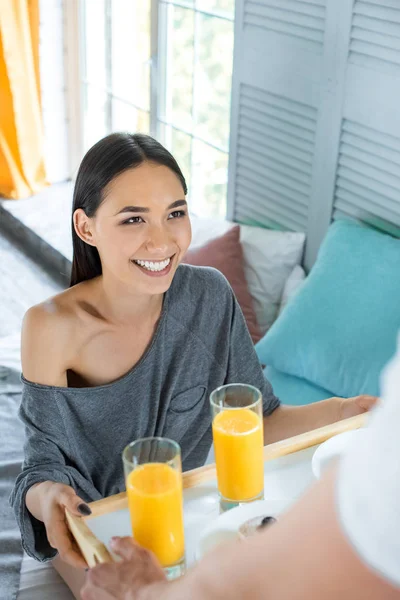 Parcial vista de hombre traído desayuno a sonriente asiático novia en cama en casa - foto de stock
