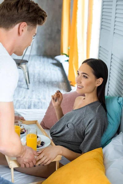 Parcial vista de hombre traído desayuno a sonriente asiático novia en cama en casa - foto de stock