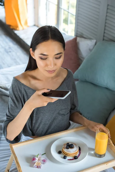 Mujer asiática tomando foto de desayuno en bandeja de madera en el teléfono inteligente en el dormitorio - foto de stock