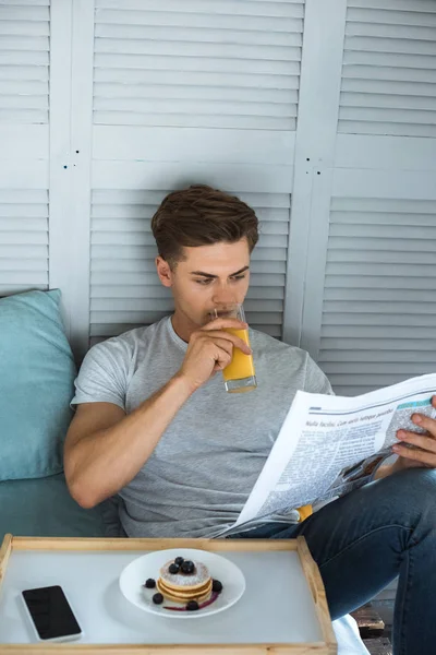 Portrait of young man drinking juice while reading newspaper in bed in morning — Stock Photo