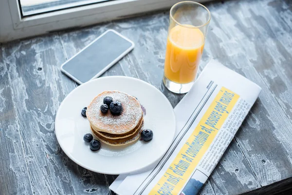 Close up view of pancakes, glass of juice, smartphone and newspaper on wooden tabletop — Stock Photo