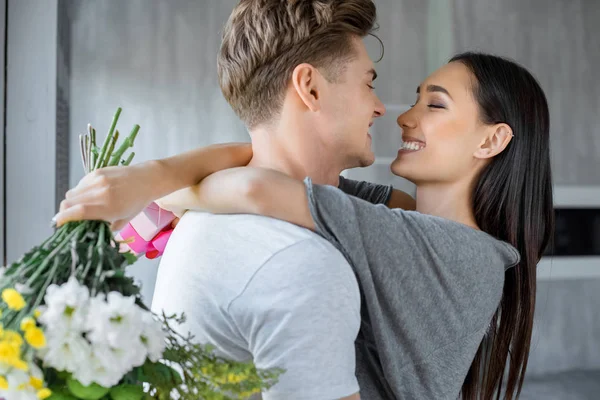 Vista laterale di felice donna asiatica con regalo e bouquet di fiori e fidanzato caucasico che si abbracciano a casa — Foto stock