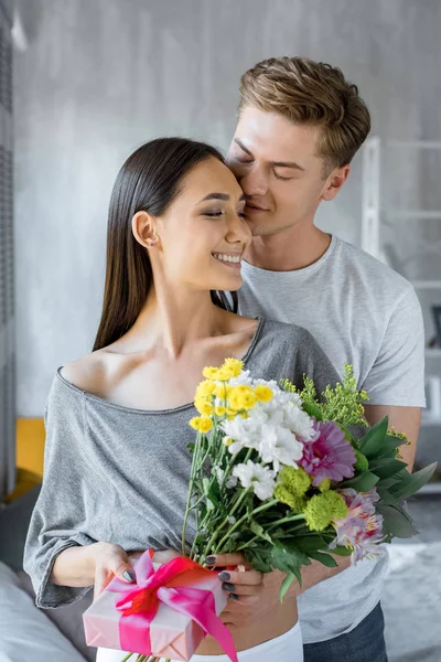 Retrato de feliz casal multiétnico com presente e buquê de flores em casa — Fotografia de Stock