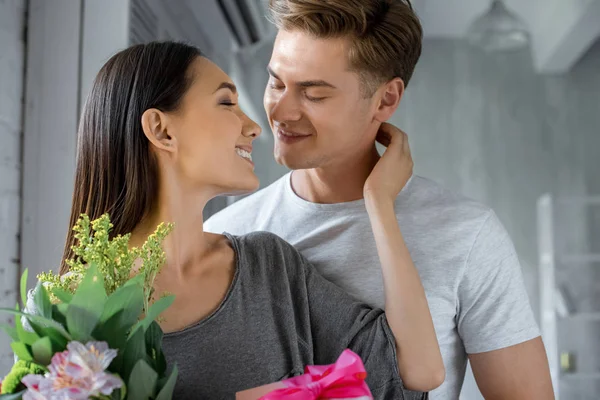 Portrait de heureuse petite amie asiatique avec présent et bouquet de fleurs regardant petit ami caucasien à la maison — Photo de stock