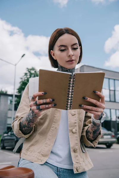 Vista de ángulo bajo de la mujer tatuada con estilo leyendo libro de texto en la calle - foto de stock