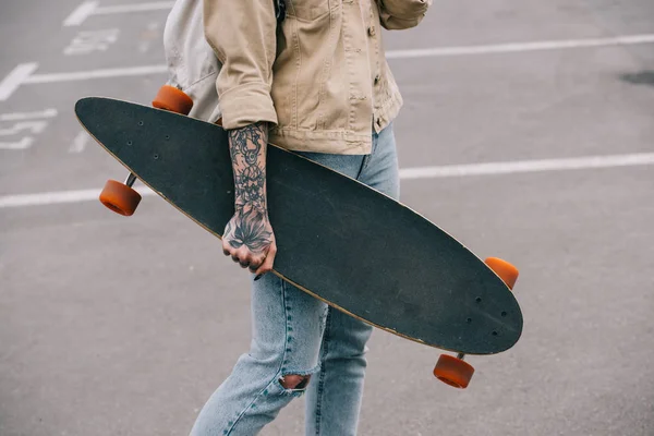 Vista parziale di elegante donna tatuata che tiene longboard al parcheggio — Foto stock