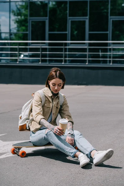 Татуированная женщина сидит на скейтборде с бумажной чашкой кофе — стоковое фото