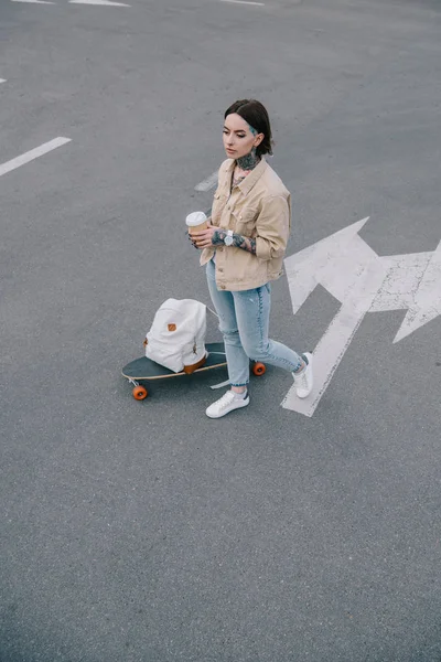 Visão de alto ângulo da mulher tatuada com xícara de café em pé no estacionamento perto de skate e mochila — Fotografia de Stock