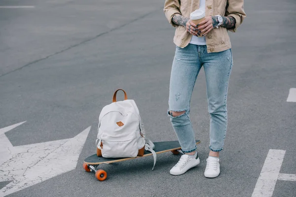 Abgeschnittenes Bild einer tätowierten stylischen Frau mit Einwegbecher Kaffee, die neben Skateboard und Rucksack auf einem Parkplatz steht — Stockfoto