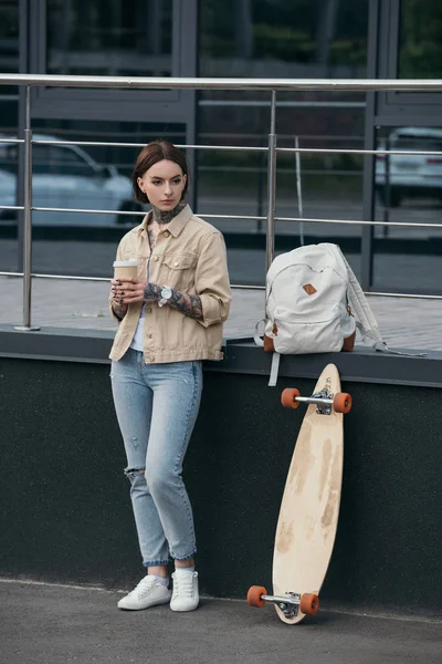 Mujer tatuada con estilo sosteniendo taza de café y de pie cerca de monopatín en la calle - foto de stock