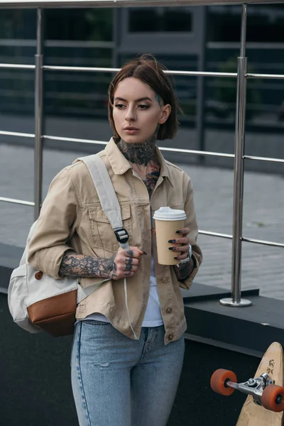 Mujer tatuada con mochila sosteniendo taza de café y de pie cerca de monopatín en la calle - foto de stock