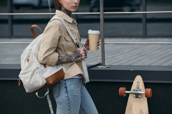 Image recadrée de femme tatouée avec sac à dos tenant tasse de café et debout près de planche à roulettes à la rue — Photo de stock