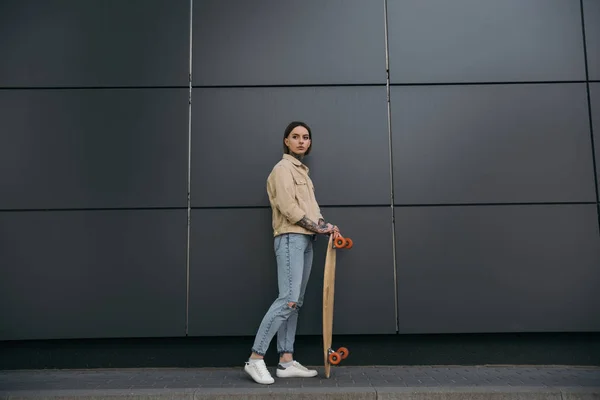 Стильная женщина с татуировками, стоящая со скейтбордом у черной стены — стоковое фото
