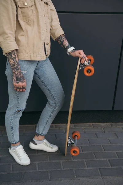 Immagine ritagliata di donna tatuata in piedi con skateboard contro muro nero — Foto stock