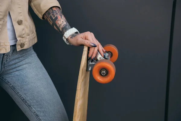 Cropped image of tattooed woman holding skateboard near black wall — Stock Photo