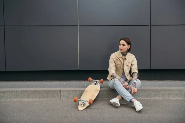 Donna elegante tatuata seduta vicino allo skateboard in strada — Foto stock